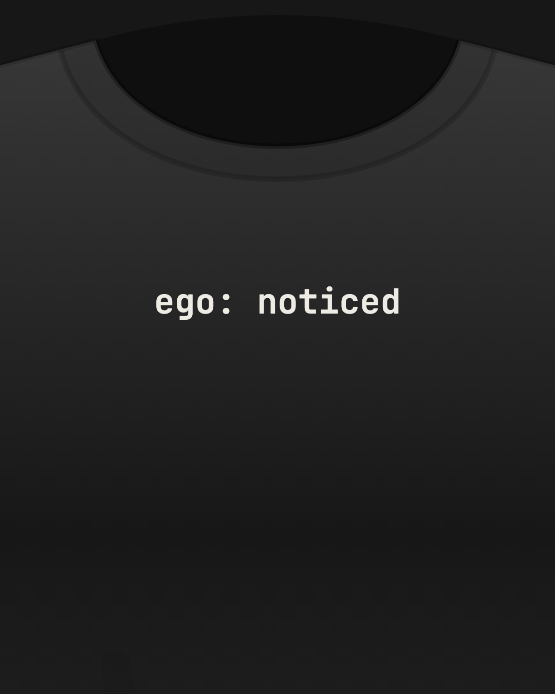 Close-up of a black tee with “ego: noticed” printed small in off-white on the chest